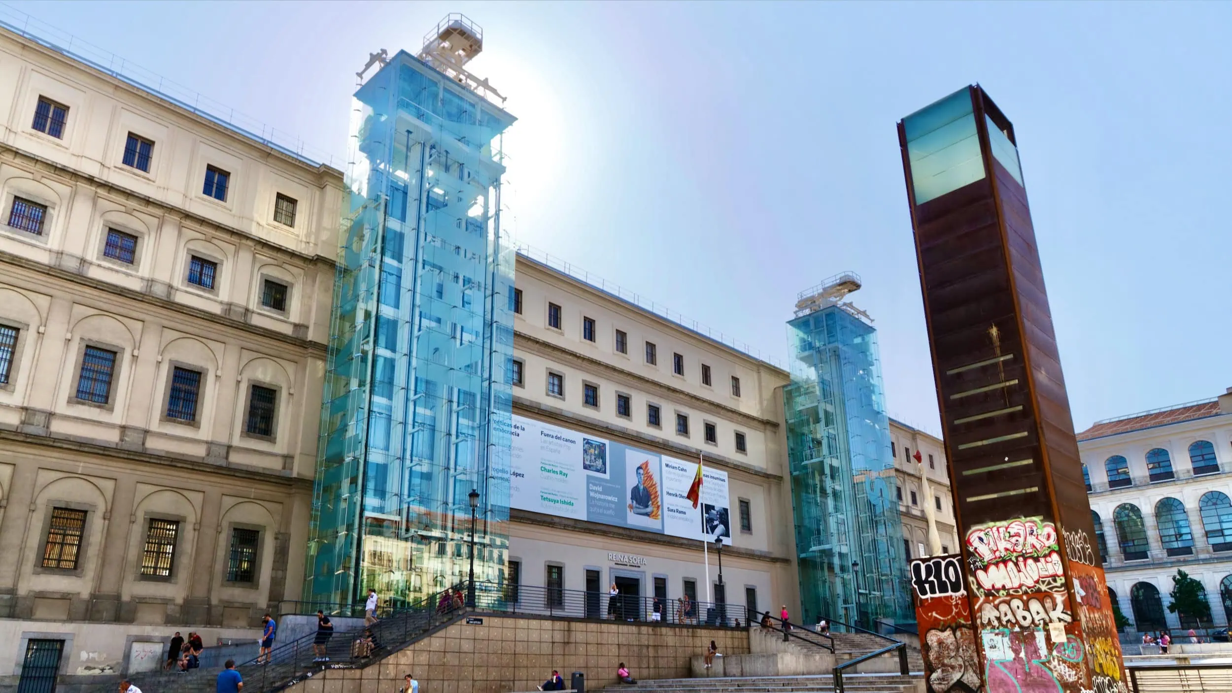 Hoofdentree van het Reina Sofía Museum met het achttiende-eeuwse Sabatini-gebouw en de moderne glazen liften rond de binnenplaats, waar bezoekers ontspannen verblijven.