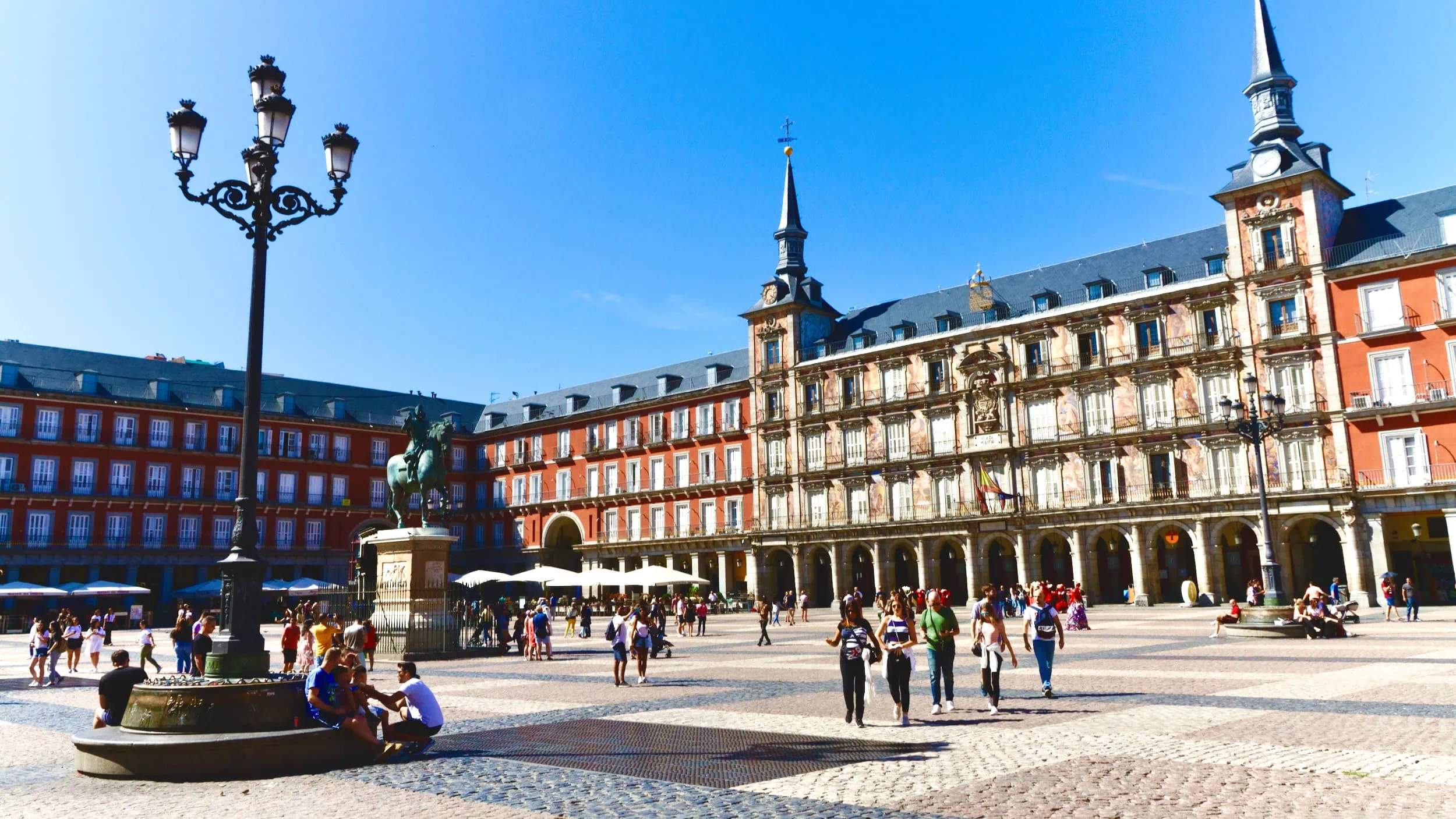 Plaza Mayor in Madrid met de Casa de la Panadería en haar kleurrijke fresco’s, onder een heldere blauwe lucht, terwijl bezoekers ontspannen wandelen en verblijven rond het plein.